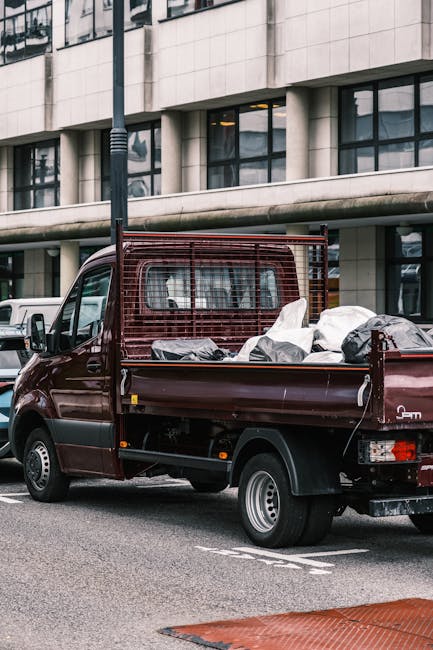 A small burgundy flatbed truck parked on an urban street in front of a modern office building with large glass windows and a tiled facade. The truck's cargo area is filled with various waste materials
